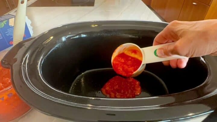 White measuring cup dumps tomato sauce into an empty slow cooker.