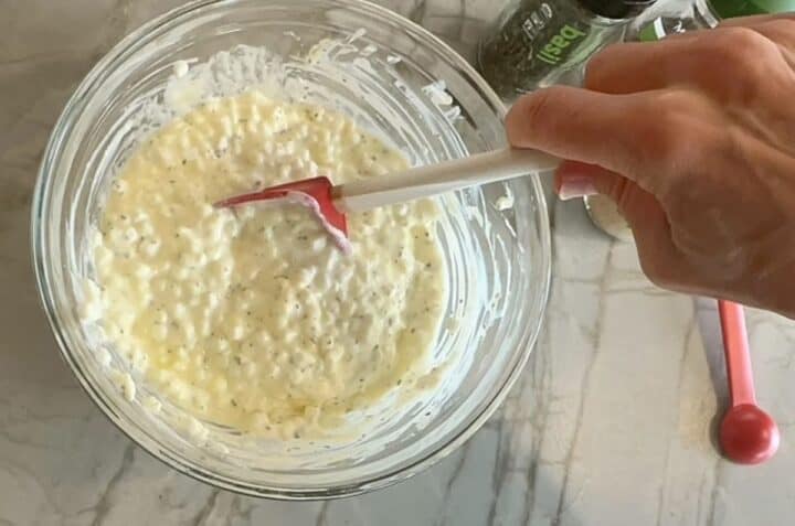 Stirring dried herbs into a clear bowl holding white cottage cheese.
