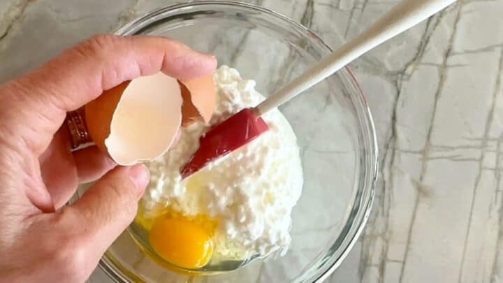 Clear bowl holding cottage cheese with a raw egg cracked into it. Brown shell held above bowl.