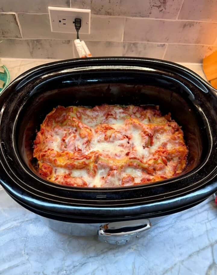 Ruffly edges of lasagna noodles poke up from melted cheese in the black liner of a slow cooker.