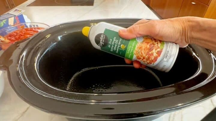 Can of non-stick olive oil spray held in front of a shiny, black slow cooker liner.