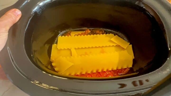 Yellow uncooked lasagna noodles top tomato sauce in black slow cooker liner.
