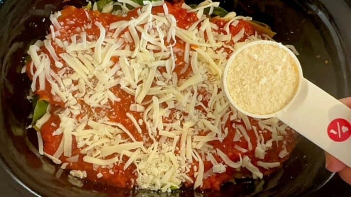White cup of grated off-white Parmesan cheese hovers over uncooked Slow Cooker Lasagna, ready to sprinkle over red sauce and white shredded cheese.