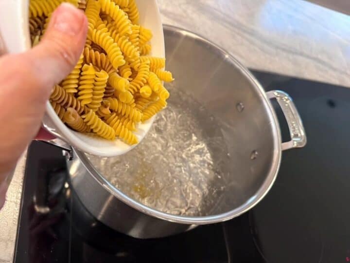 An aluminum pot of boiling water awaits golden colored curly pasta being poured into it.