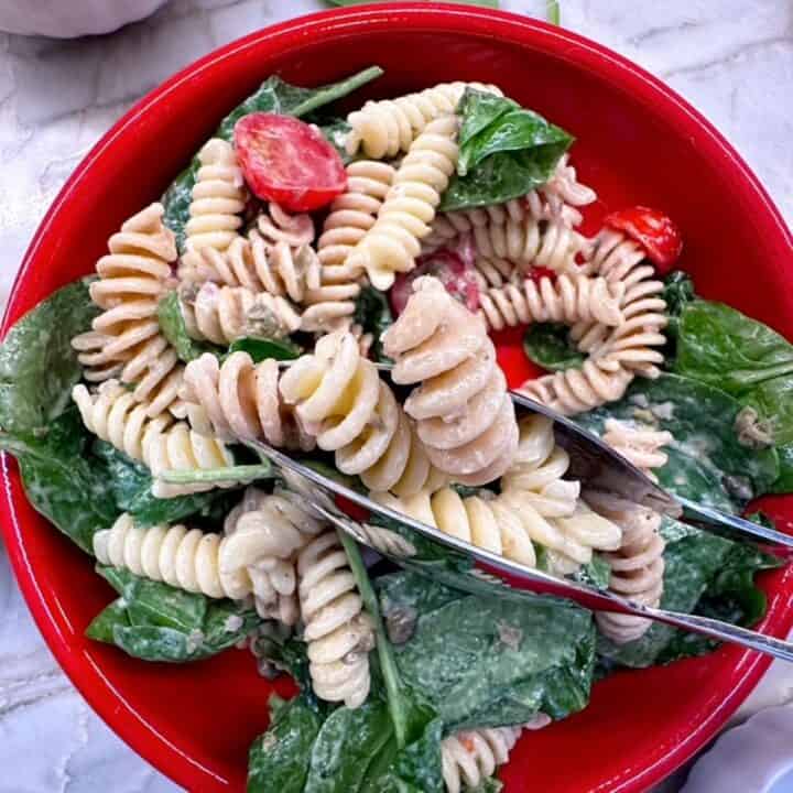 Silver tongs reach into a round red bowl for corkscrew pasta pieces and spinach in a creamy Greek yogurt dressing.