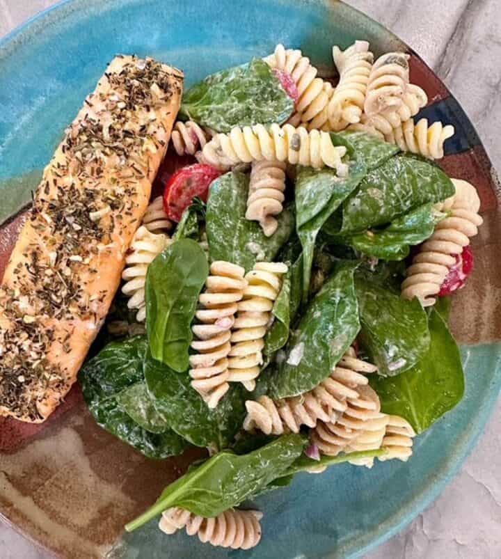 Turquoise pottery plate holds a grilled seasoned salmon fillet and corkscrew pasta salad with spinach leaves tossed in a creamy Greek yogurt dressing.