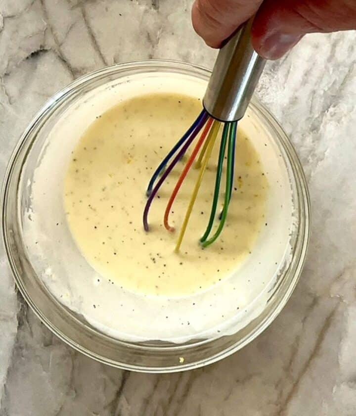 A multicolored silicone whisk emulsifes Greek yogurt dressing with lemon into a creamy light yellow mixture in a clear bowl.