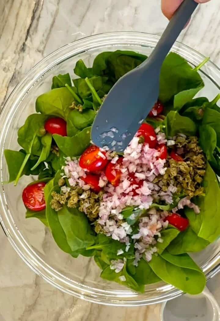 Green baby spinach, cherry tomatoes, minced shallots, and more ingredients in a clear bowl ready to be dressed.