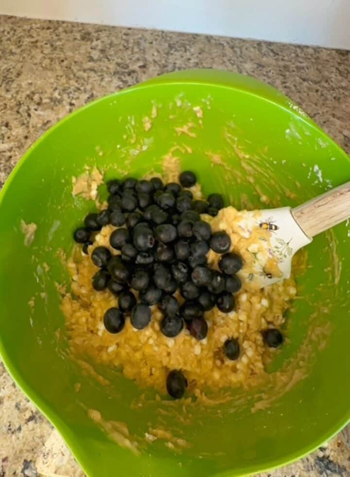 Dark violet blueberries sit atop muffin batter in a lime green bowl ready to be mixed by the inserted spatula.