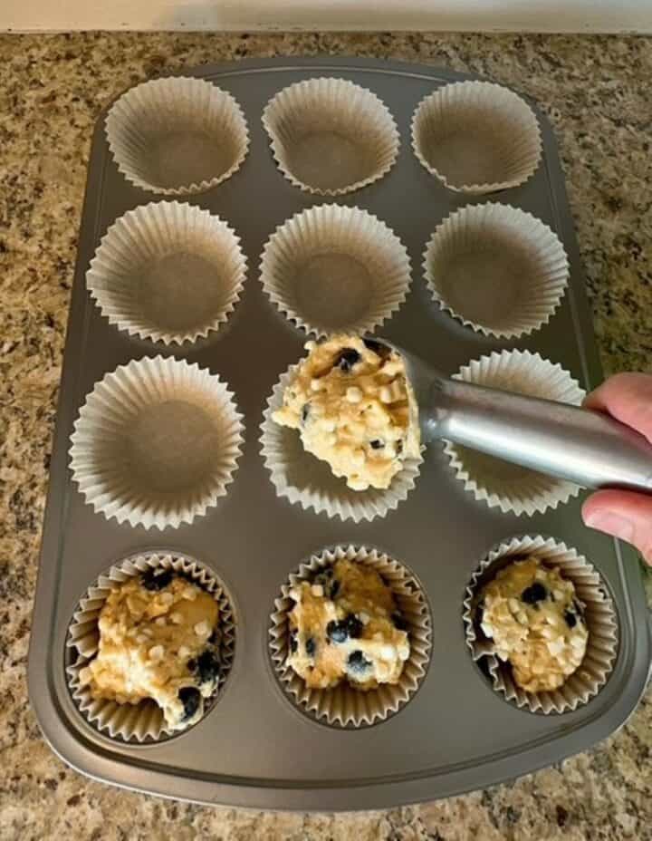 Metal ice cream scoop portions blueberry muffin batter into semi-opaque liners in a muffin tin.