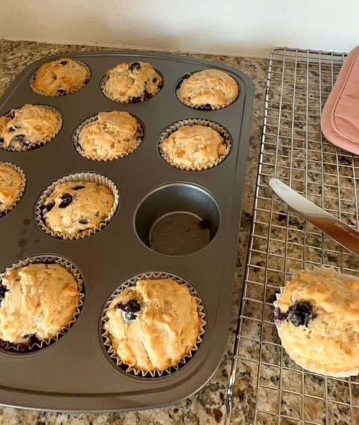 One blueberry muffin sits on a cooling rack next to a brown tin full of fresh baked muffins.