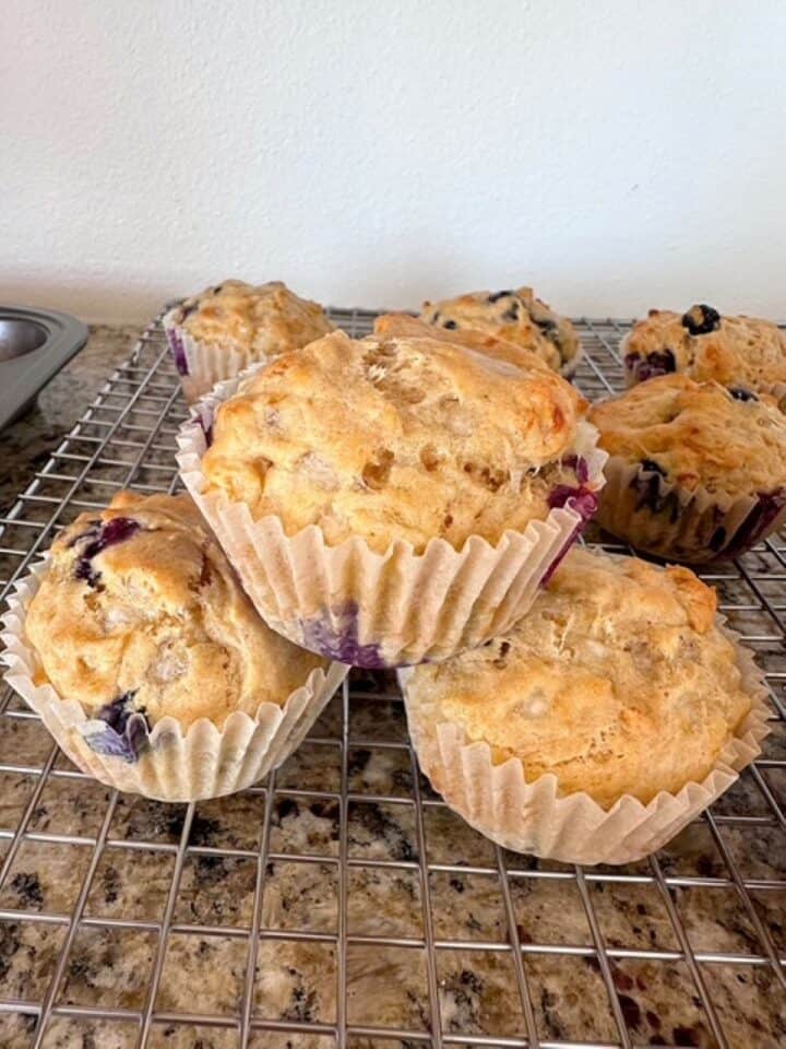 Three blueberry muffins are stacked like a triangle on a cooling rack by more blueberry muffins.