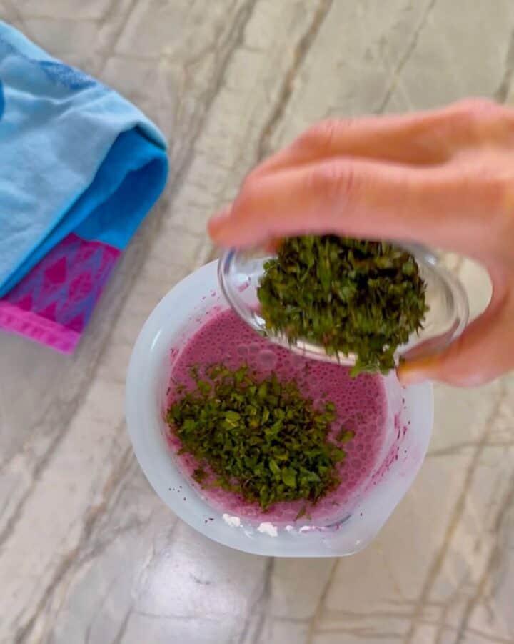 Pouring green chopped mint from a small, clear bowl into an opaque container of purplish blueberry-yogurt puree.