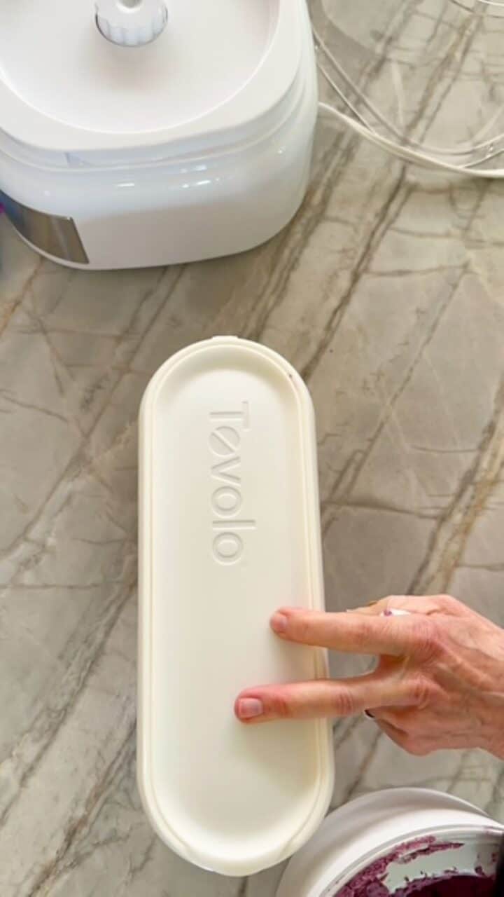 Two fingers tap a long white oval container full of blueberry froyo.