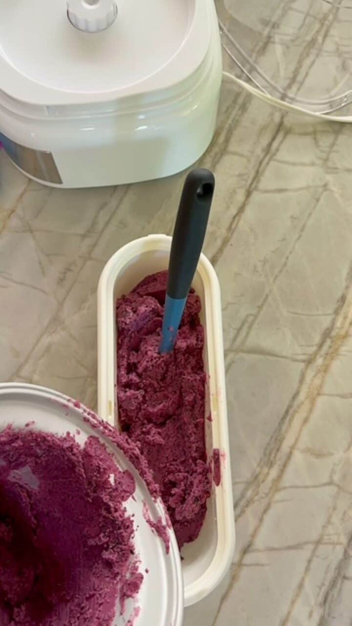 Transferring purplish blueberry froyo from ice cream maker bowl into white insulated container.