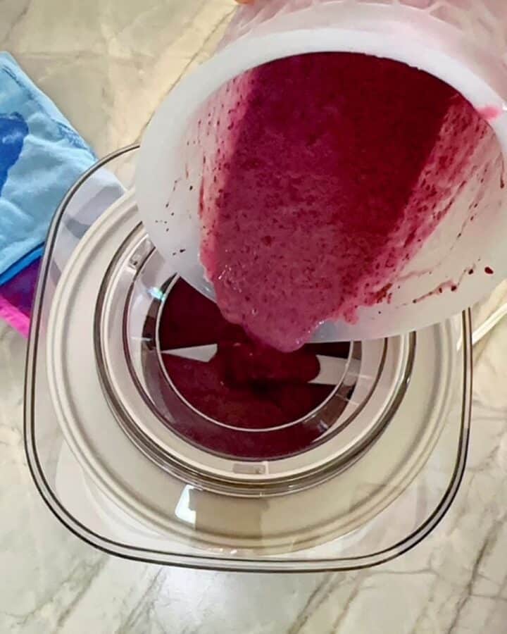 Pouring purplish blueberry-yogurt puree into the circular opening on top of a white ice cream maker.
