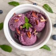 Scoops of purple blueberry frozen yogurt with mint fill a round white ramekin.
