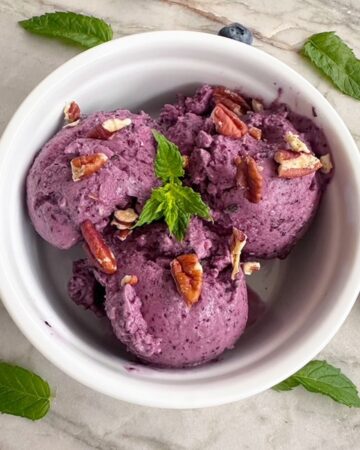 Scoops of purple blueberry frozen yogurt with mint fill a round white ramekin.