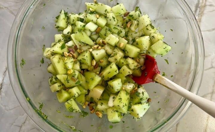 Salad of honeydew cubes with flecks of mint and a red spatula for serving out of a clear bowl.