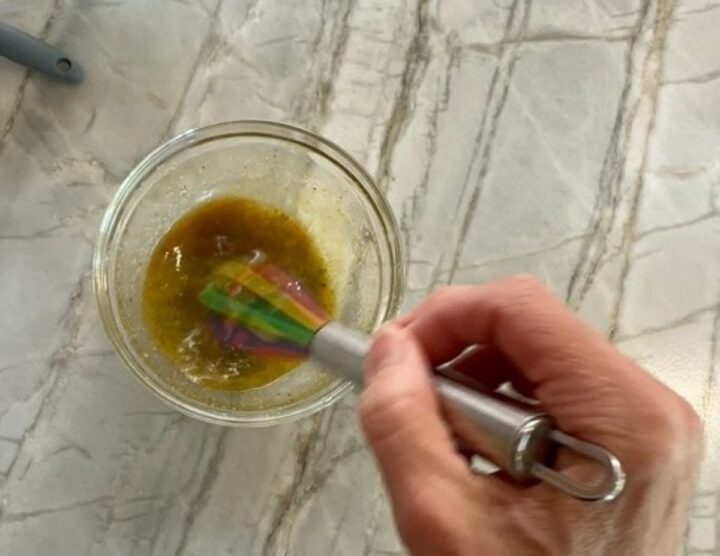Beating light brown dressing with a silver whisk in a small, clear bowl.