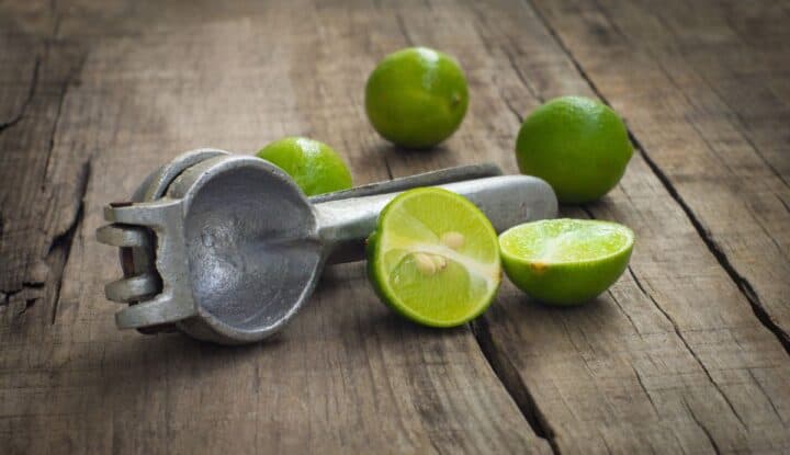 Metal squeezer with cut and whole green limes on a wooden board.