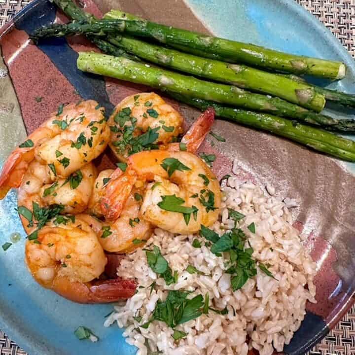 Pink shrimp sprinkled with green parsley are nestled together with brown rice and green asparagus spears on a pottery plate streaked with turquoise and brown.