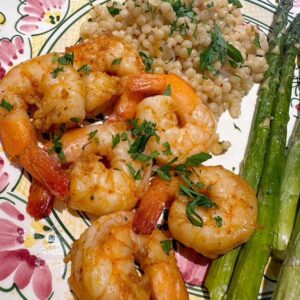 Closeup of large pink shrimp sprinkled with chopped dark green parsley sitting next to cooked green asparagus and pearl couscous.