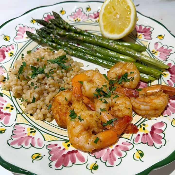 Hand-painted plate with pink, yellow, and green designs holds big pink shrimp, green asparagus spears and beige pearl couscous.