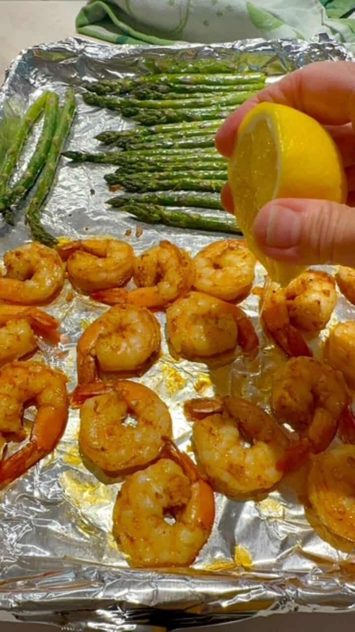 Squeezing half a lemon over cooked, large pink shrimp and green asparagus on a foil-lined sheet pan.