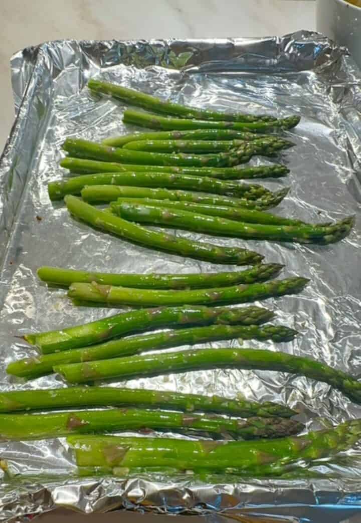 Green, oil-slicked asparagus spears on a shiny, foil-lined sheet pan.