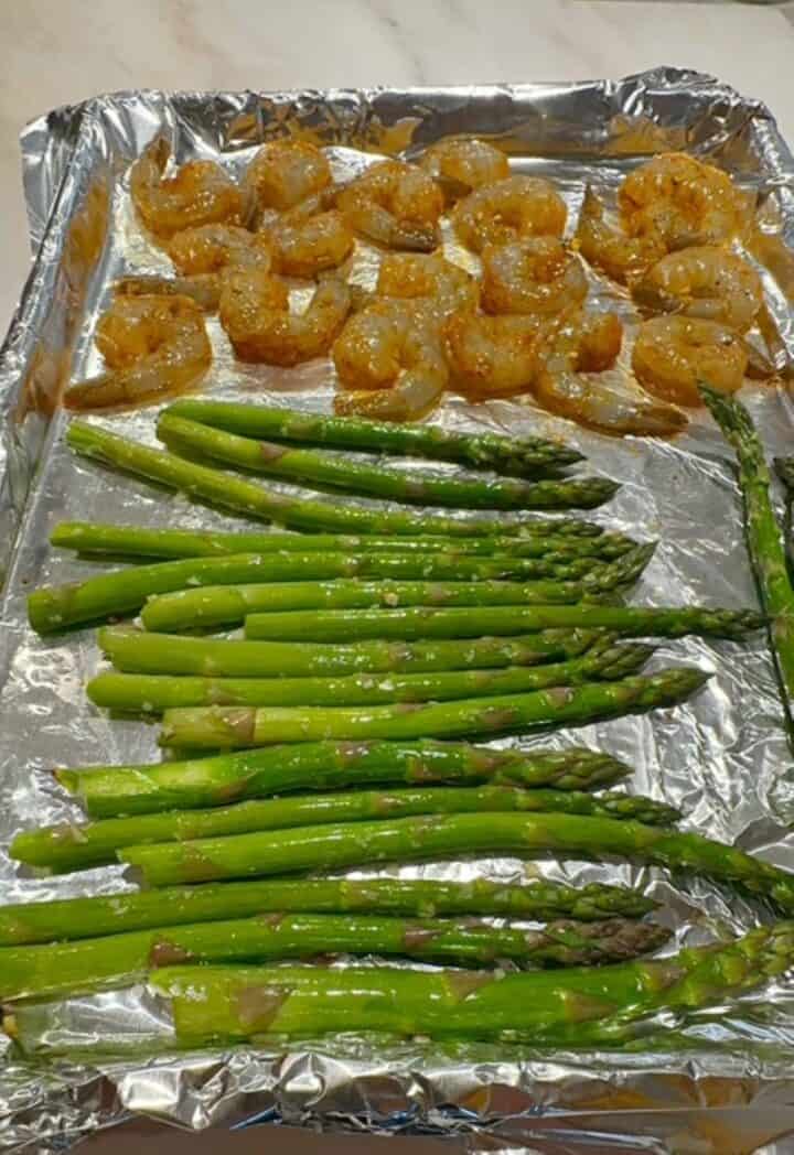 Shiny, foil-lined sheet pan with green asparagus spears on one side and pinkish seasoned, raw shrimp on the other.