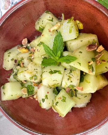 Light green honeydew cubes flecked with dark green mint and topped with a mint spring in a medium brown pottery bowl.