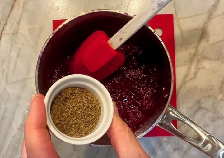 Chia powder is added to saucepan of cooked blackberries from a small white pinch bowl.