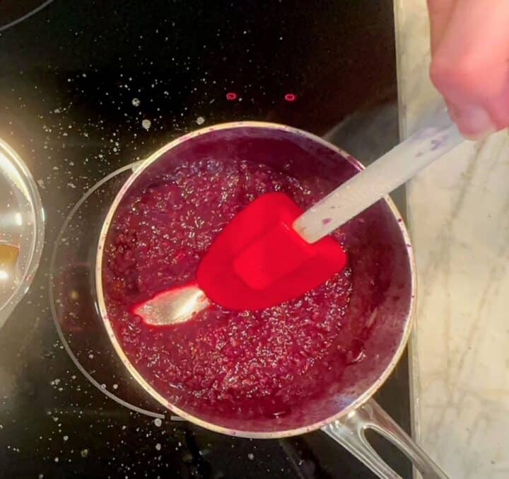 A red spatula is dragged through magenta cooked berries in a small saucepan on a black cooktop.