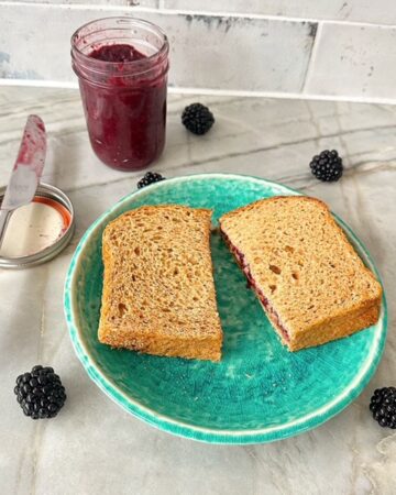 Open jar of red-purple blackberry jam is behind a green plate holding a pb&j sandwich cut in half.