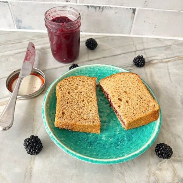 Open jar of red-purple blackberry jam is behind a green plate holding a pb&j sandwich cut in half.