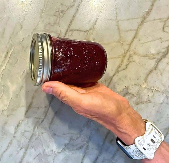 A small jar of blackberry jam is presented in the palm of a hand on a gray marble countertop.