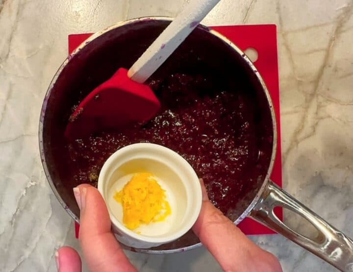 Yellow lemon zest is added to saucepan of cooked blackberries from a small white pinch bowl.