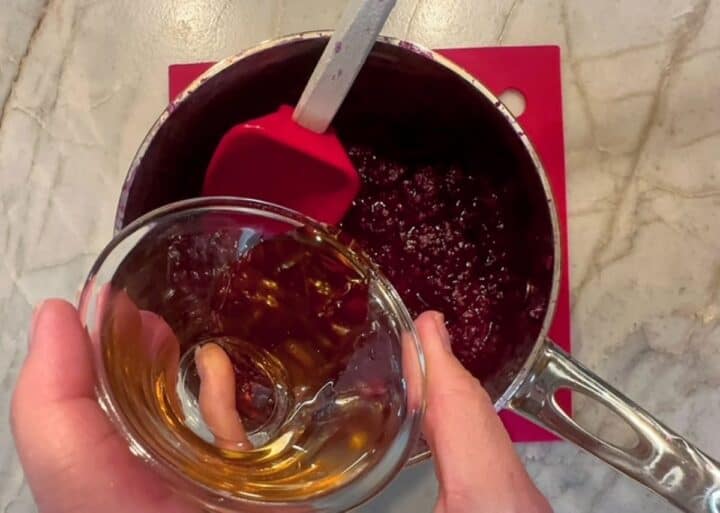 Hand pours amber maple syrup into saucepan of cooked berries.