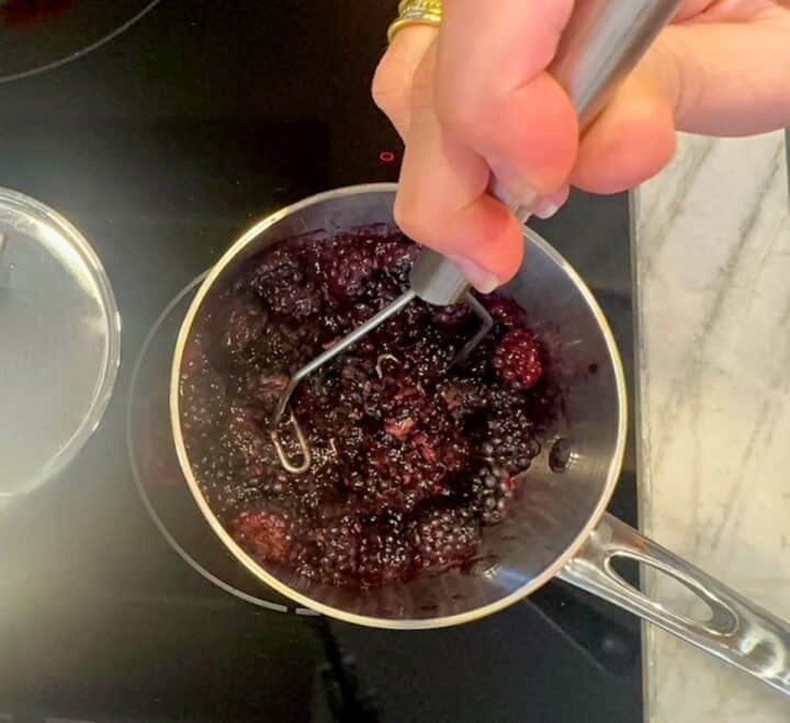 Mashing blackberries in a small saucepan on a black cooktop with a silver potato masher.