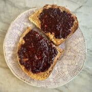 A piece of wheat toast covered with nut butter and purple blackberry chia jam sits on a pottery plate.