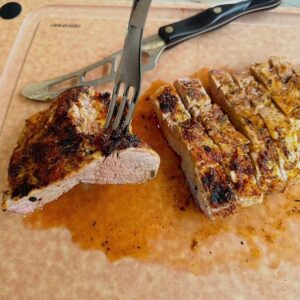 Brown cutting board holds a grilled, sliced pork tenderloin with a serving fork sticking into it.