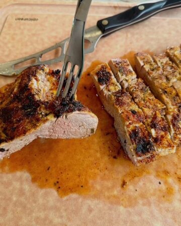 Brown cutting board holds a grilled, sliced pork tenderloin with a serving fork sticking into it.
