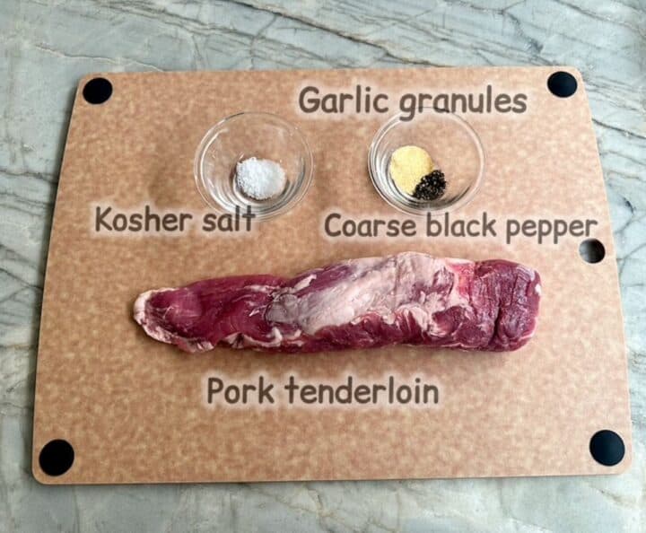 A raw, red and skinny pork tenderloin sits on a light brown cutting board with salt, garlic granules, and black pepper.
