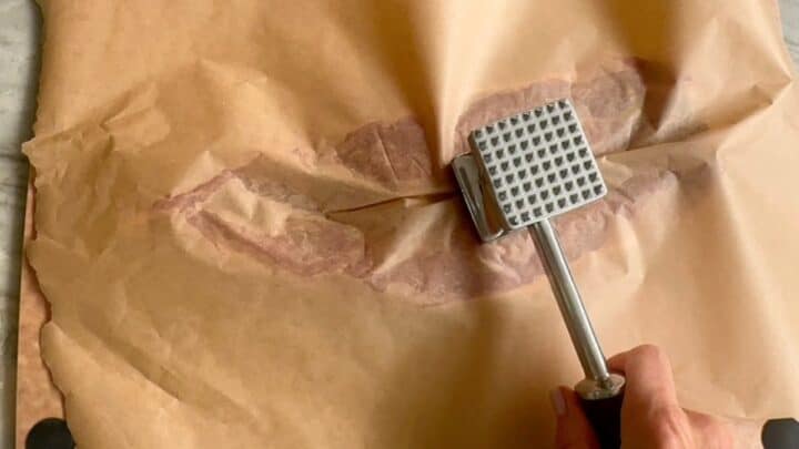 A silver meat mallet pounds on a sheet of brown wax paper over a raw pork tenderloin.