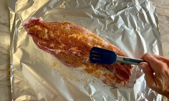 A seasoned raw, butterflied pork tenderloin sits on a piece of shiny foil with tongs on top of it.