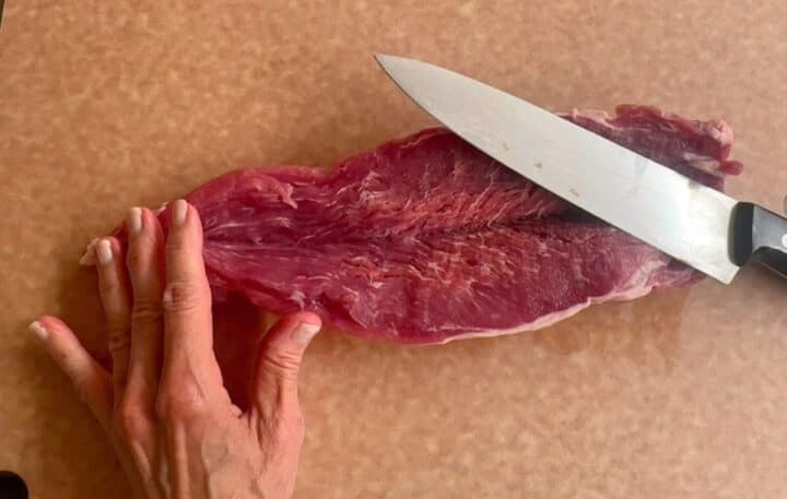 Light brown cutting board holds a raw, red pork tender that has been cut down the middle.