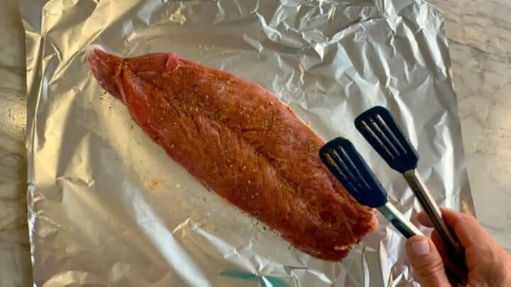 A raw, red, butterflied pork tenderloin sits flat on a piece of shiny foil covered with a spice seasoning.