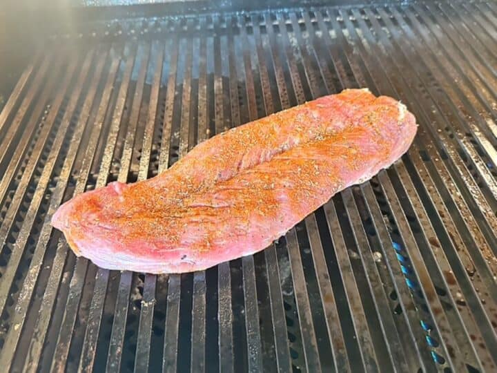A raw, pink and flat butterflied pork tender cooks on black grill grates.