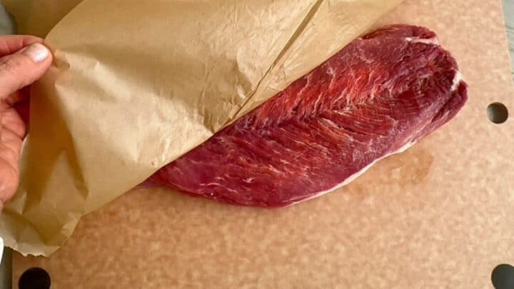 Removing brown waxed paper to reveal a flattened, raw pork tenderloin on a cutting board.